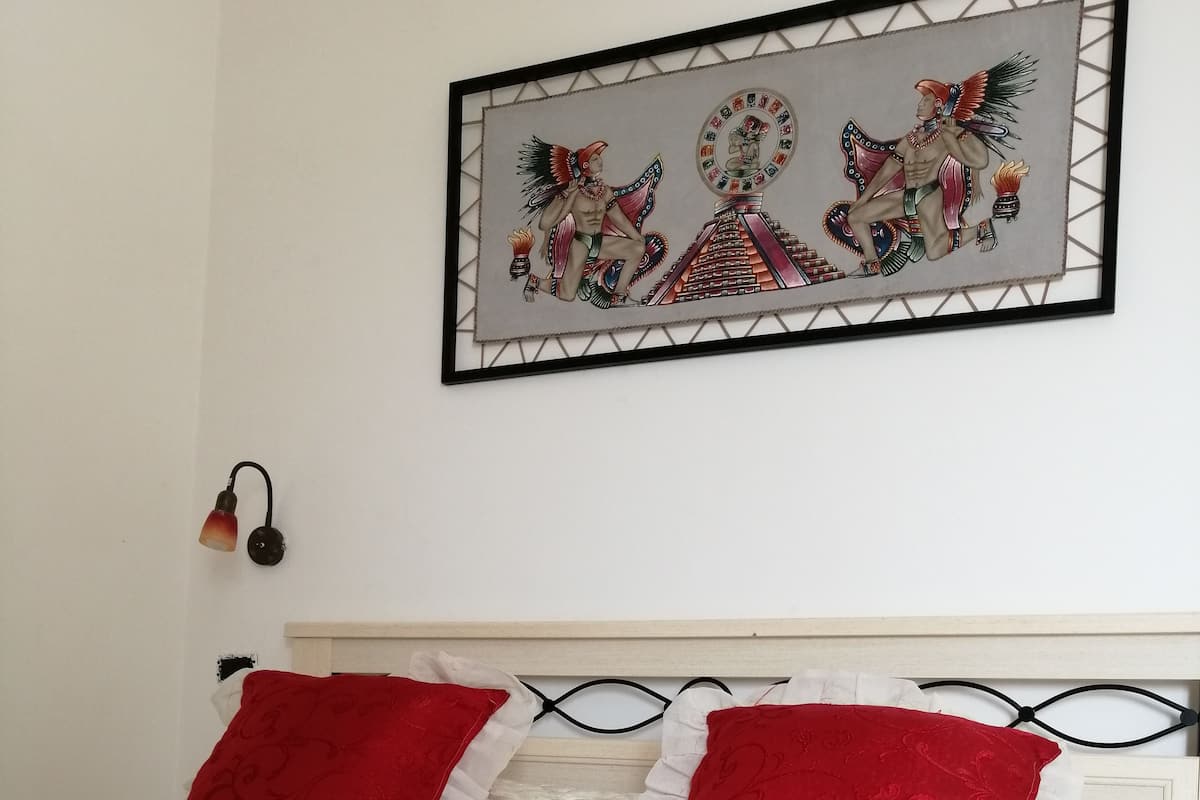 A neatly made bed with red and white pillows, a tray with pastries and chocolates, and a framed artwork above the bed.