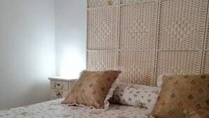 Un lit bien fait, avec une literie à motifs floraux, un coussin décoratif et un panier rempli de serviettes pliées, surmonté d’une fleur.