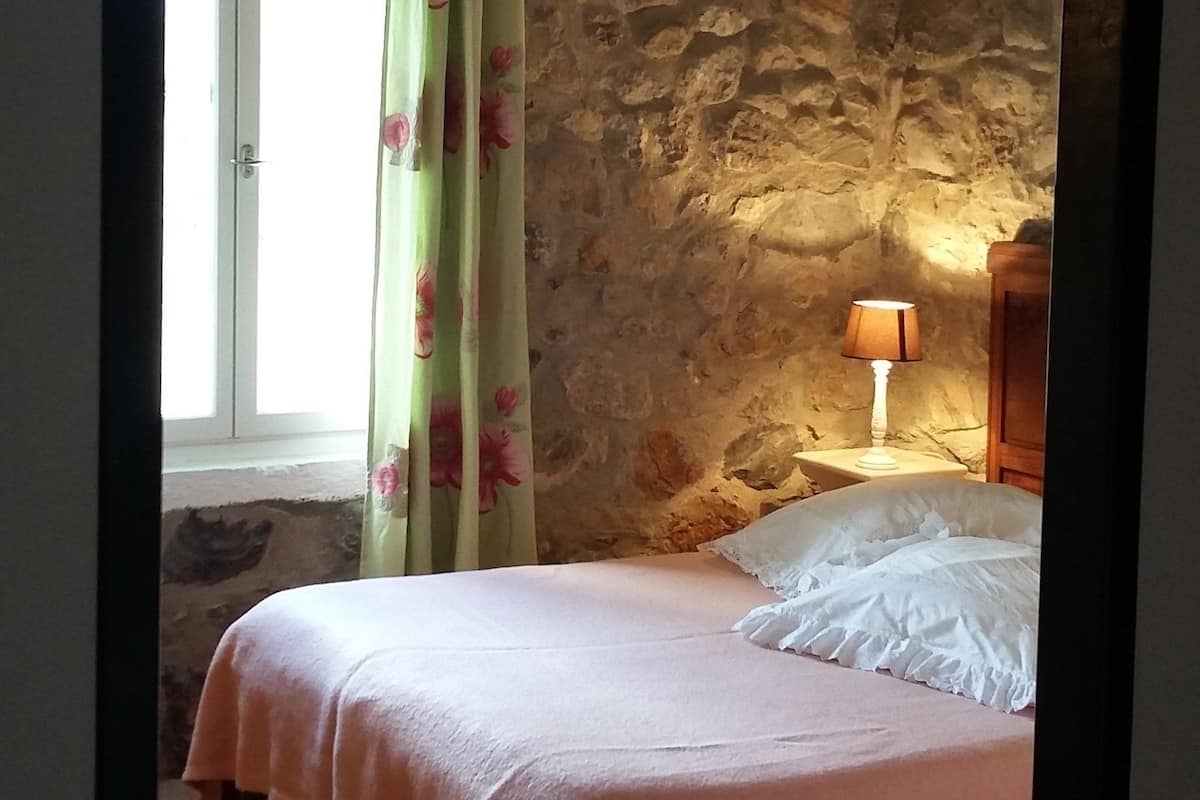 Un dormitori amb paret de pedra, llit de fusta, finestra amb cortines i una làmpada de nit.