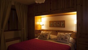 A bedroom with a wooden headboard, a red bedspread, two white pillows, a wall-mounted lamp, and a window with curtains. - Le Rosier B&B e Appartamento (Saint-Vincent)