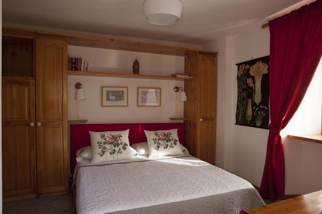 A bedroom with a bed, wooden wardrobe, shelves with books, framed pictures, and a painting.