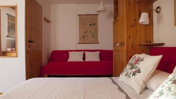 Une pièce avec un canapé rouge, un lit blanc orné d’oreillers à motifs floraux, des murs en bois et une estampe botanique suspendue au mur.