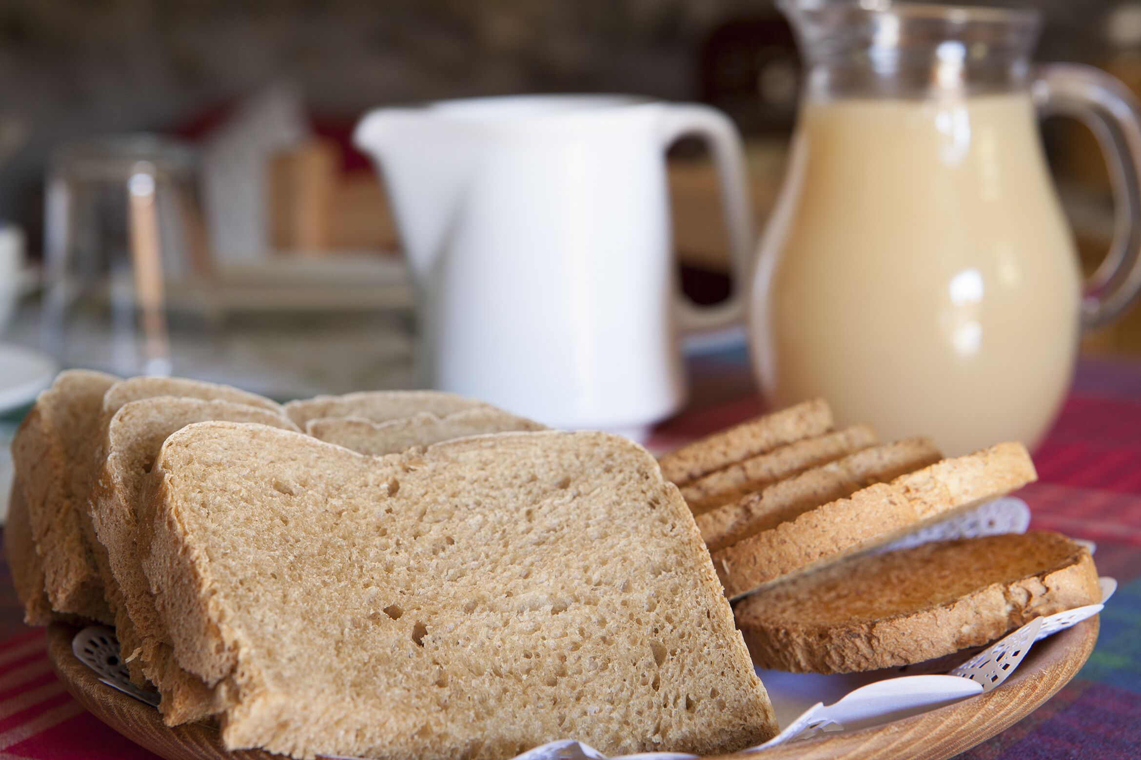 Des tranches de pain complet sur une assiette en bois, une carafe de lait et, au fond, une autre carafe blanche.