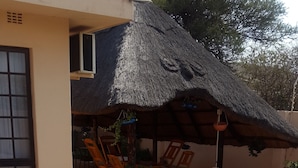 A swimming pool with a tiled edge, surrounded by a brick patio. There is a thatched-roof structure with chairs and potted plants. - Tintech Bed & Breakfast (Francistown)