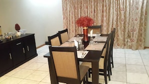 A dining area with a long table set for four, surrounded by chairs with cushioned seats. A vase with red flowers is placed on the table, and a black cabinet is visible to the left. - Tintech Bed & Breakfast (Francistown)