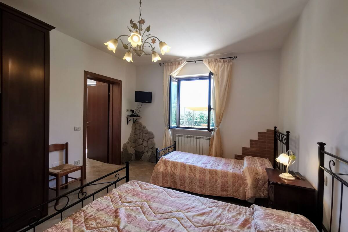 A bedroom with two beds, a stone wall, a TV, a window with curtains, and a chandelier.