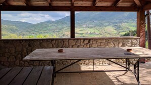 A wooden table with a stone wall backdrop, a stone floor, and a view of green hills. - Agriturismo Sole Luna (Assisi)