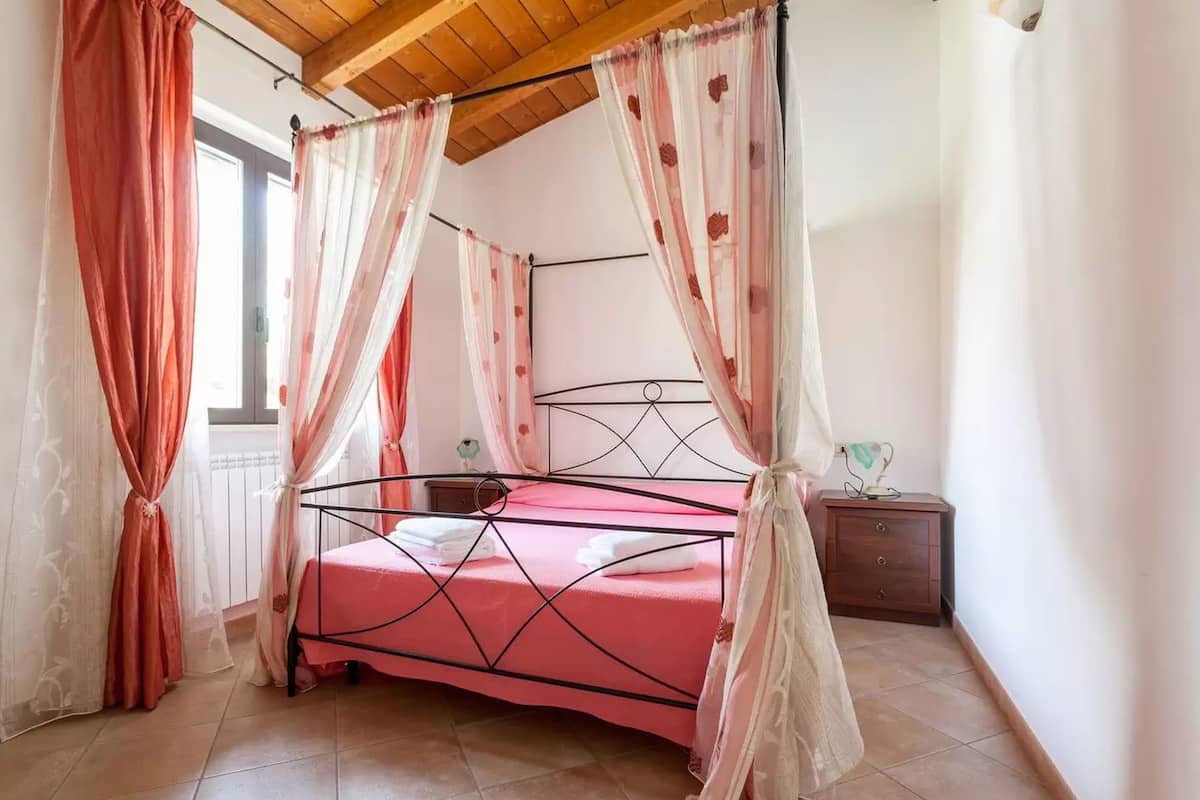 A bedroom with a four-poster bed, a wooden ceiling, a nightstand, and a window with curtains.