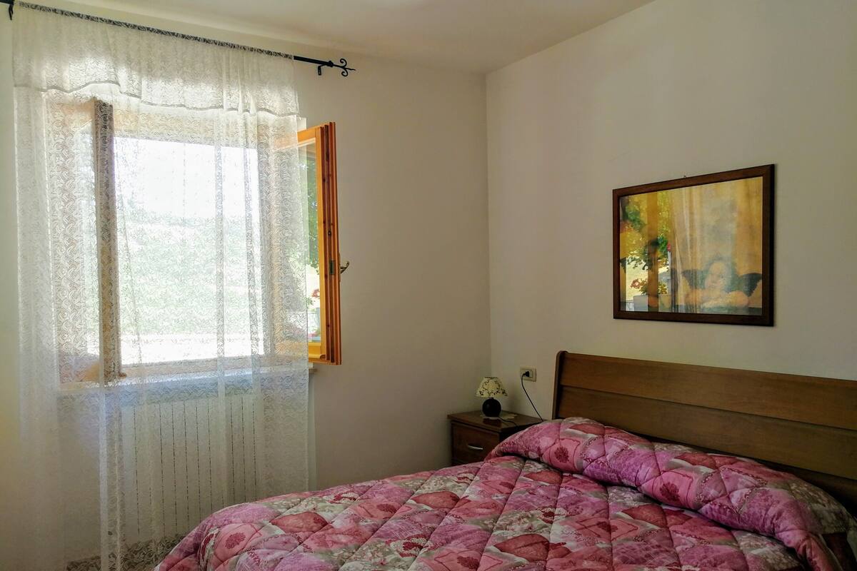 A bedroom with a bed, a nightstand, a painting, and a window with curtains.