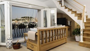 Sala de estar com sofá de madeira, televisão, planta em vaso e vista para um porto através de uma porta de vidro.