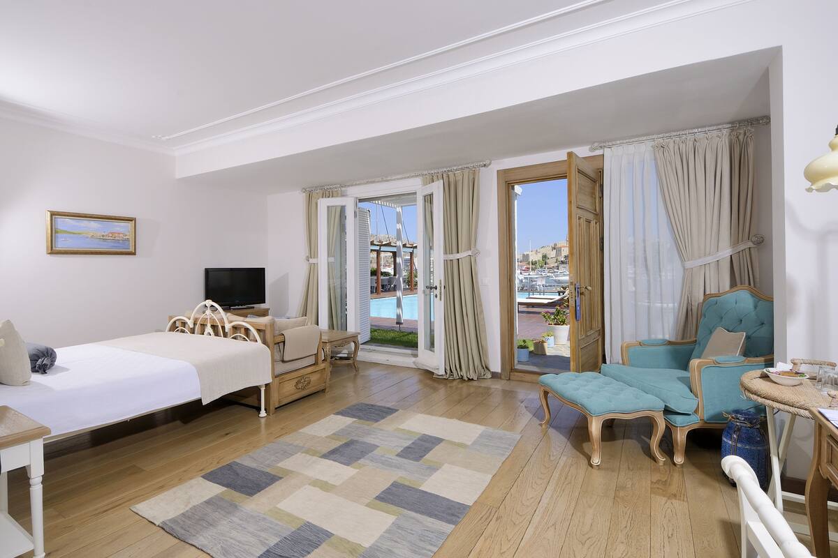 Une chambre d’hôtel équipée d’un lit, d’une télévision, d’un canapé, d’un fauteuil, d’une petite table et offrant une vue sur un port de plaisance.