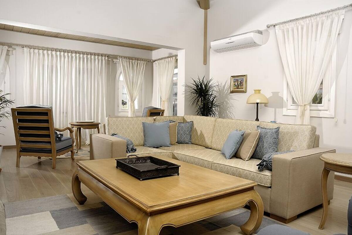 Un salon avec un canapé beige, une table basse en bois, une chaise en bois et une petite table d’appoint.
