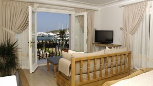 Quarto de hotel com sofá de madeira, televisão sobre suporte de madeira e varanda com vista para barcos e vegetação.