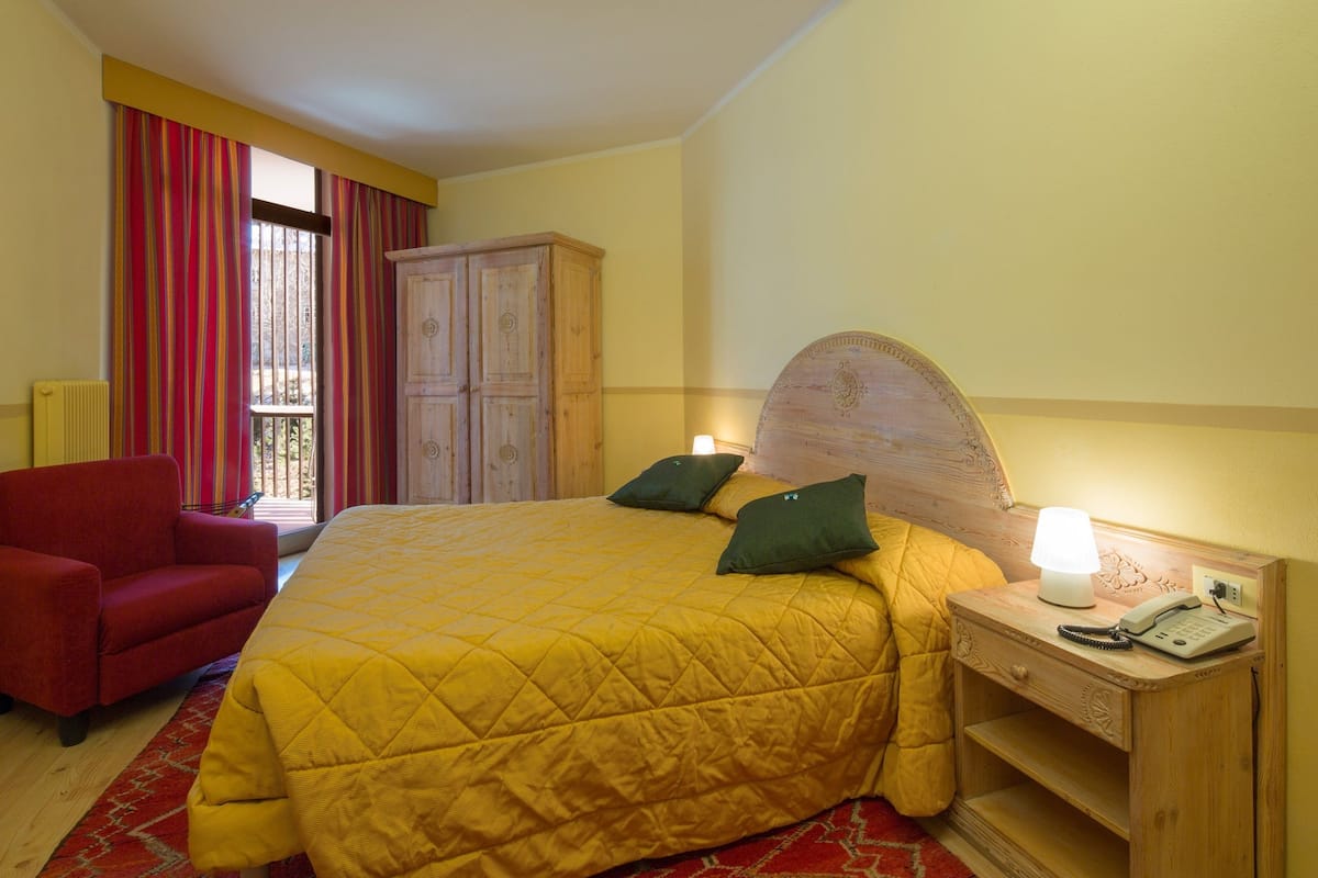 A bedroom with a large bed, a wooden nightstand with a lamp, a red armchair, and a window with striped curtains.