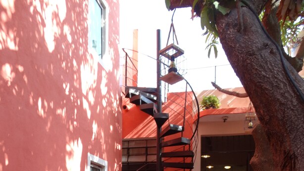 A patio with a wooden table and chairs, a spiral staircase, and a red building facade. - Casa San Juan (Mérida)