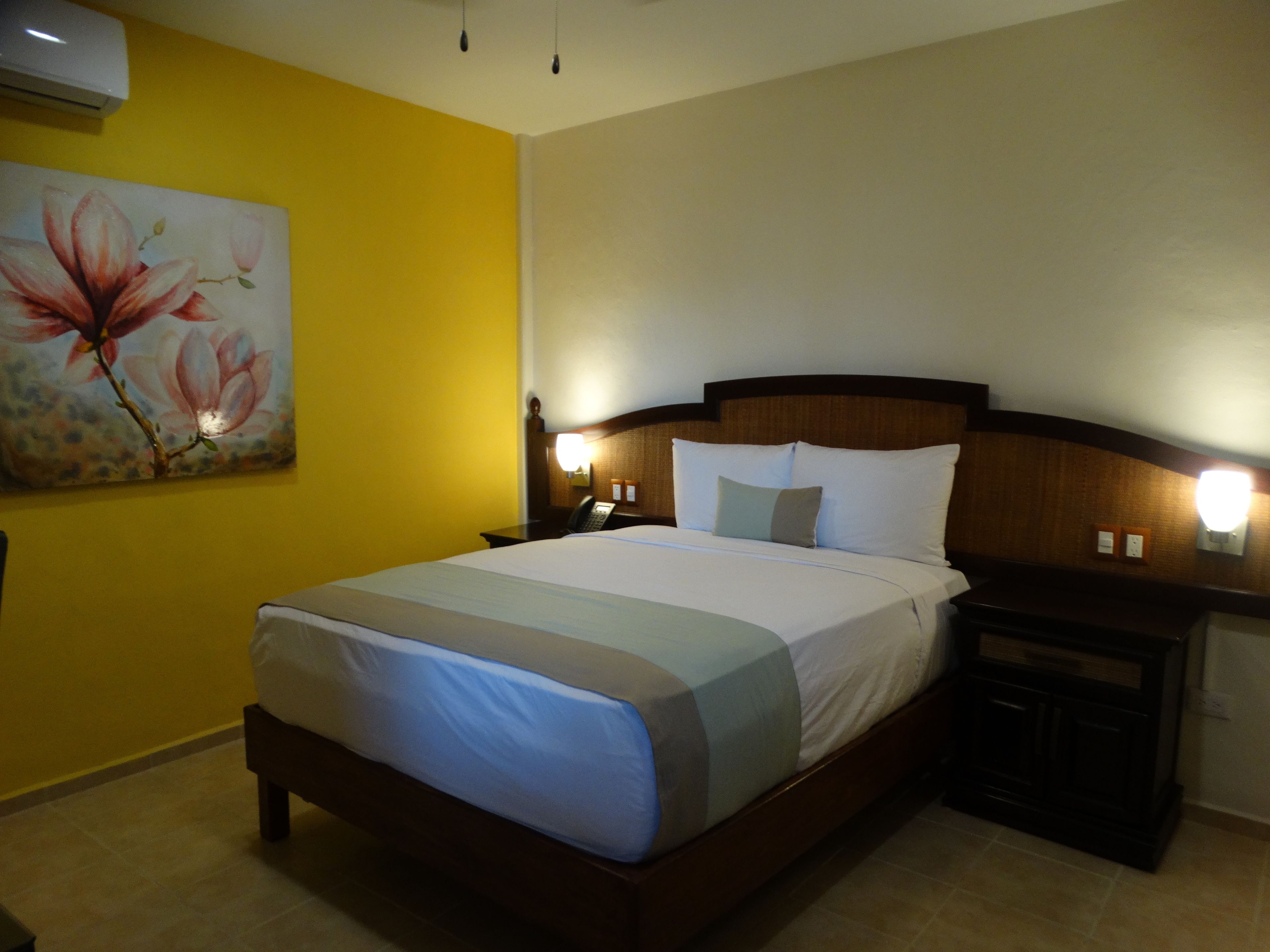 A hotel room with a bed, nightstands, a painting, and a wall-mounted air conditioner.