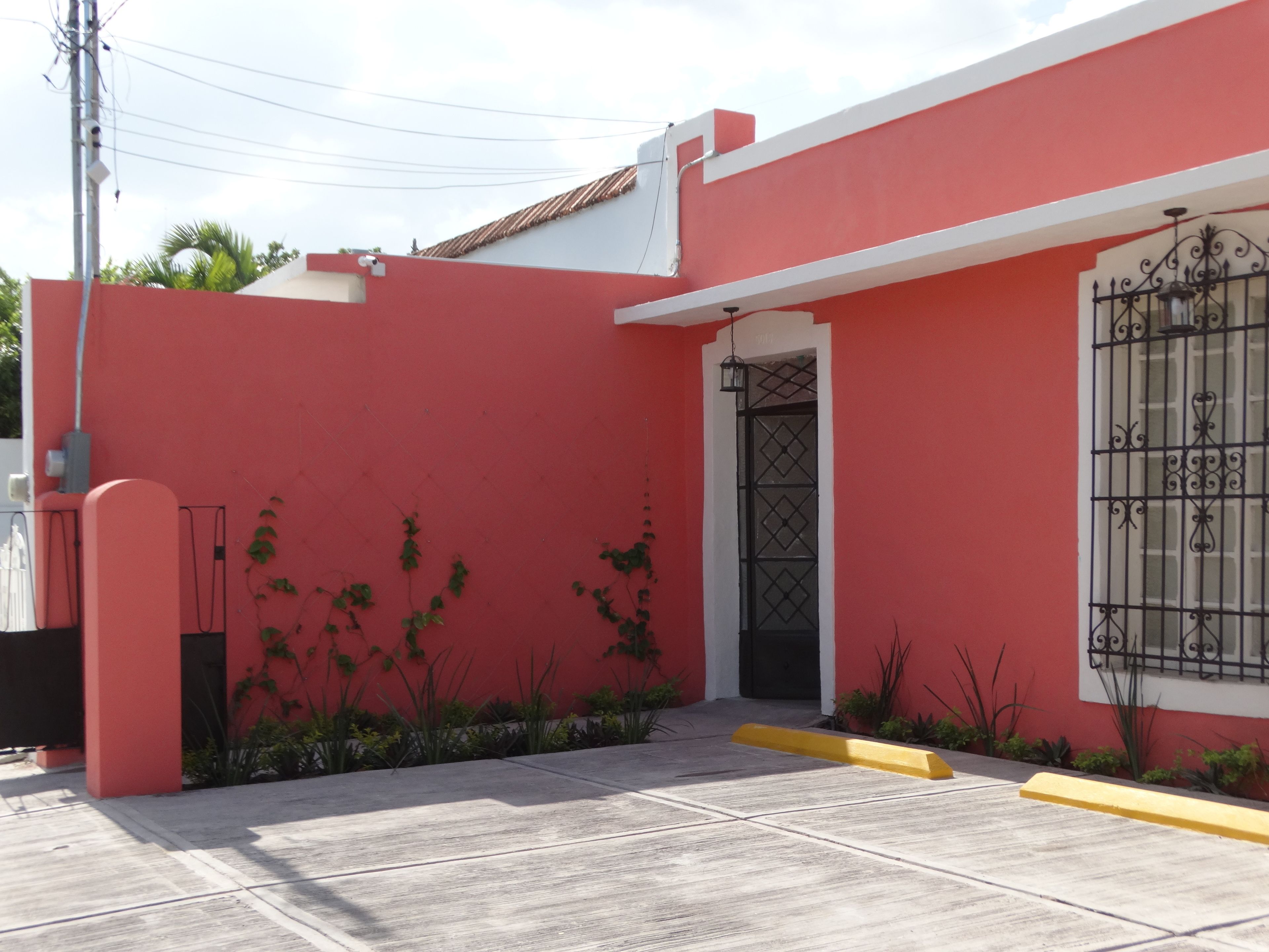 Un edificio rosso con ingresso chiuso da cancello, una finestra con inferriate decorative e un piccolo giardino con piante.