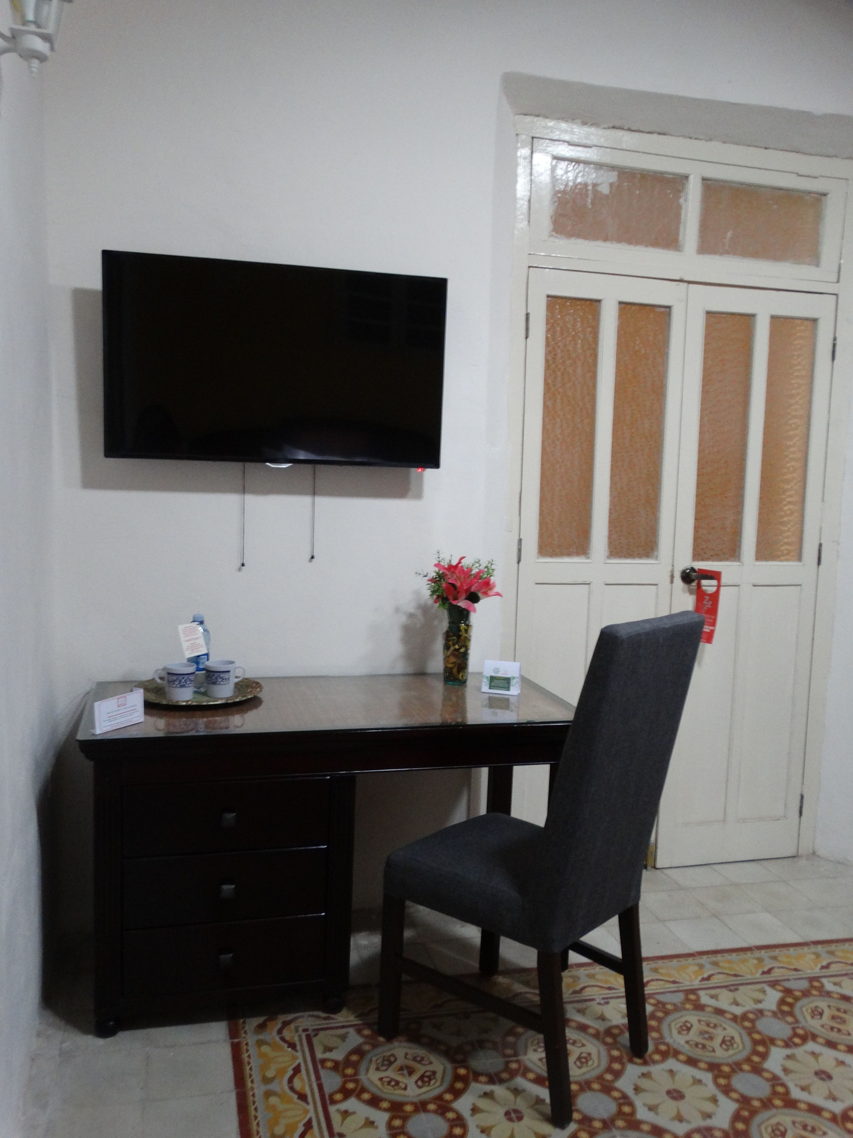 A room with a dark wooden desk, a chair, a flat-screen TV mounted on the wall, a small vase with flowers, and a door with frosted glass panels.