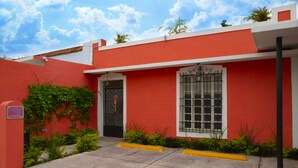 A red building with a black door and window, surrounded by greenery and a yellow curb. - Casa San Juan (Mérida)