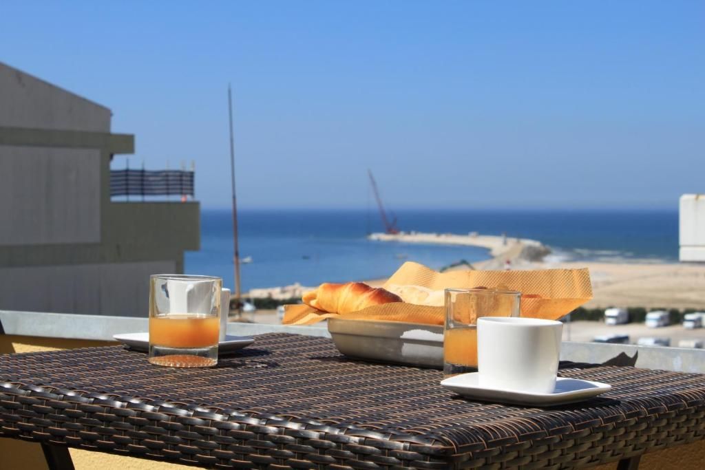 A wicker table with a glass of orange juice, a white cup, and a basket of bread on a balcony overlooking a beach.
