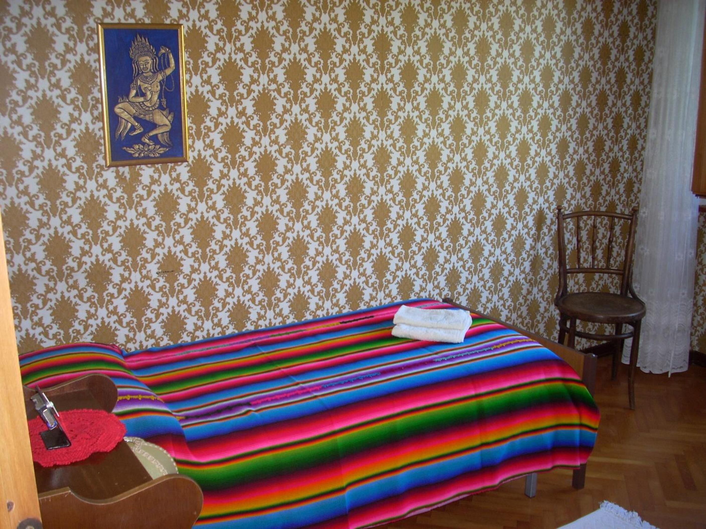 Una stanza con carta da parati decorata, un letto con coperta a righe colorate, una sedia e un quadro appeso al muro.