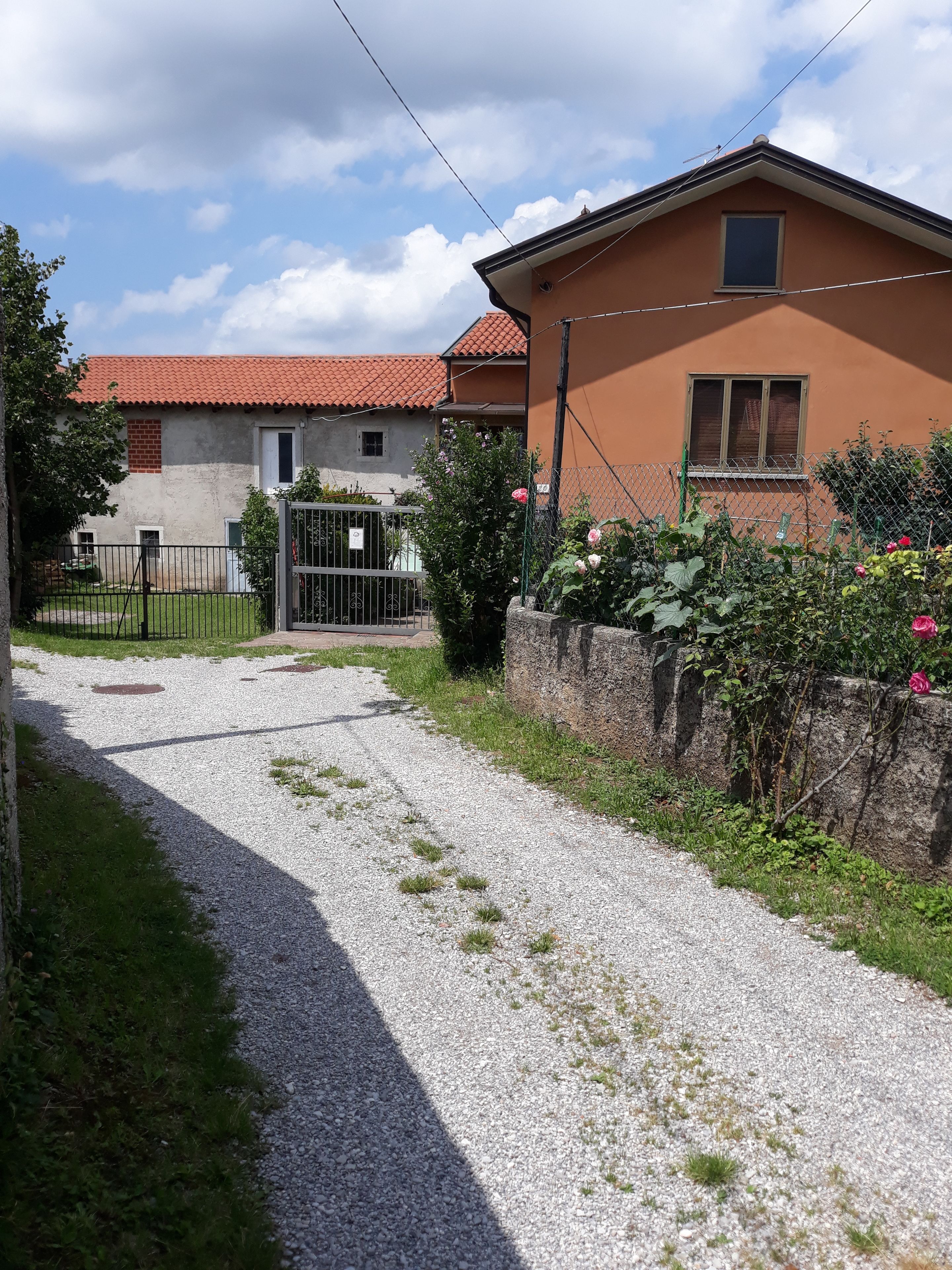 Une allée de gravier mène à une maison dont le toit est recouvert de tuiles rouges et qui est entourée d’un mur de pierres.