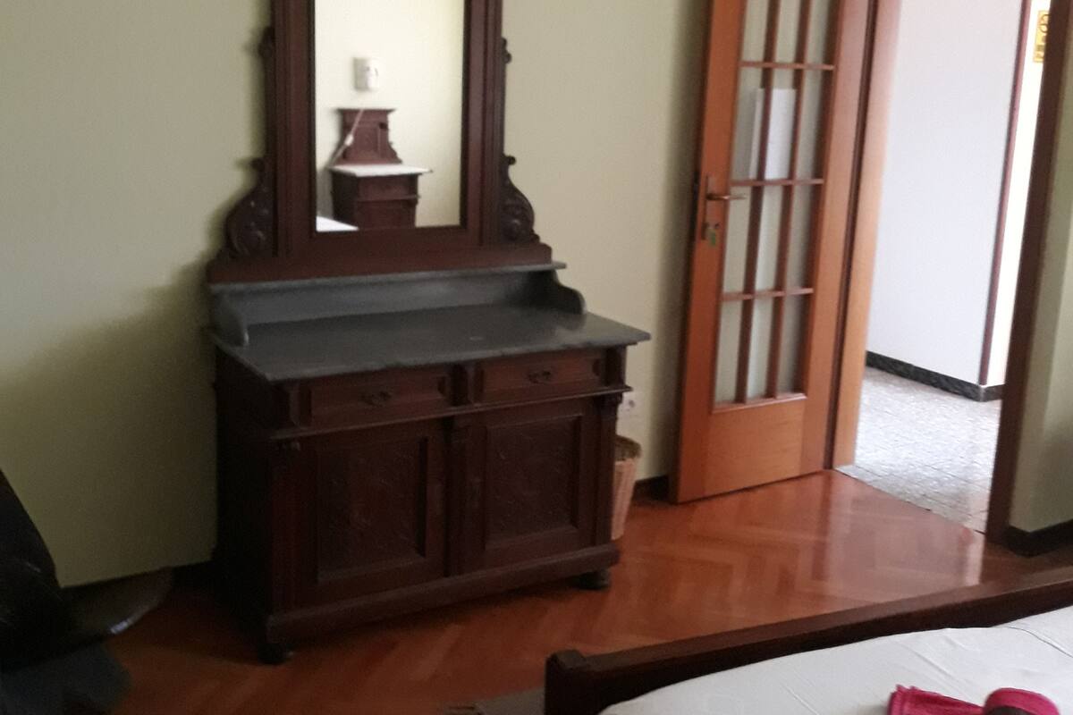 A room with a wooden dresser, a mirror, a bed with a white bedspread, and a wooden door with a glass panel.