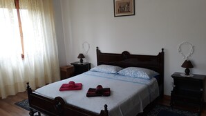 Una cama bien hecha con cabecera de madera, dos mesitas de noche con lámparas, un cuadro en la pared y una ventana con cortinas translúcidas.