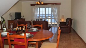 A dining area with a round table set for four, wooden chairs, a chandelier, and a view through a window with curtains. - Casa Vista Bela by Our Madeira (Calheta)