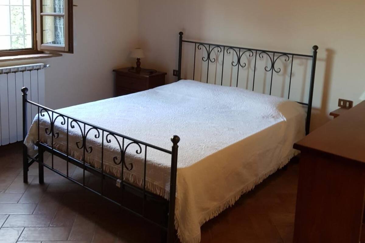 Un dormitorio con estructura de cama metálica, una ventana con vistas, un radiador y una mesita de noche pequeña.