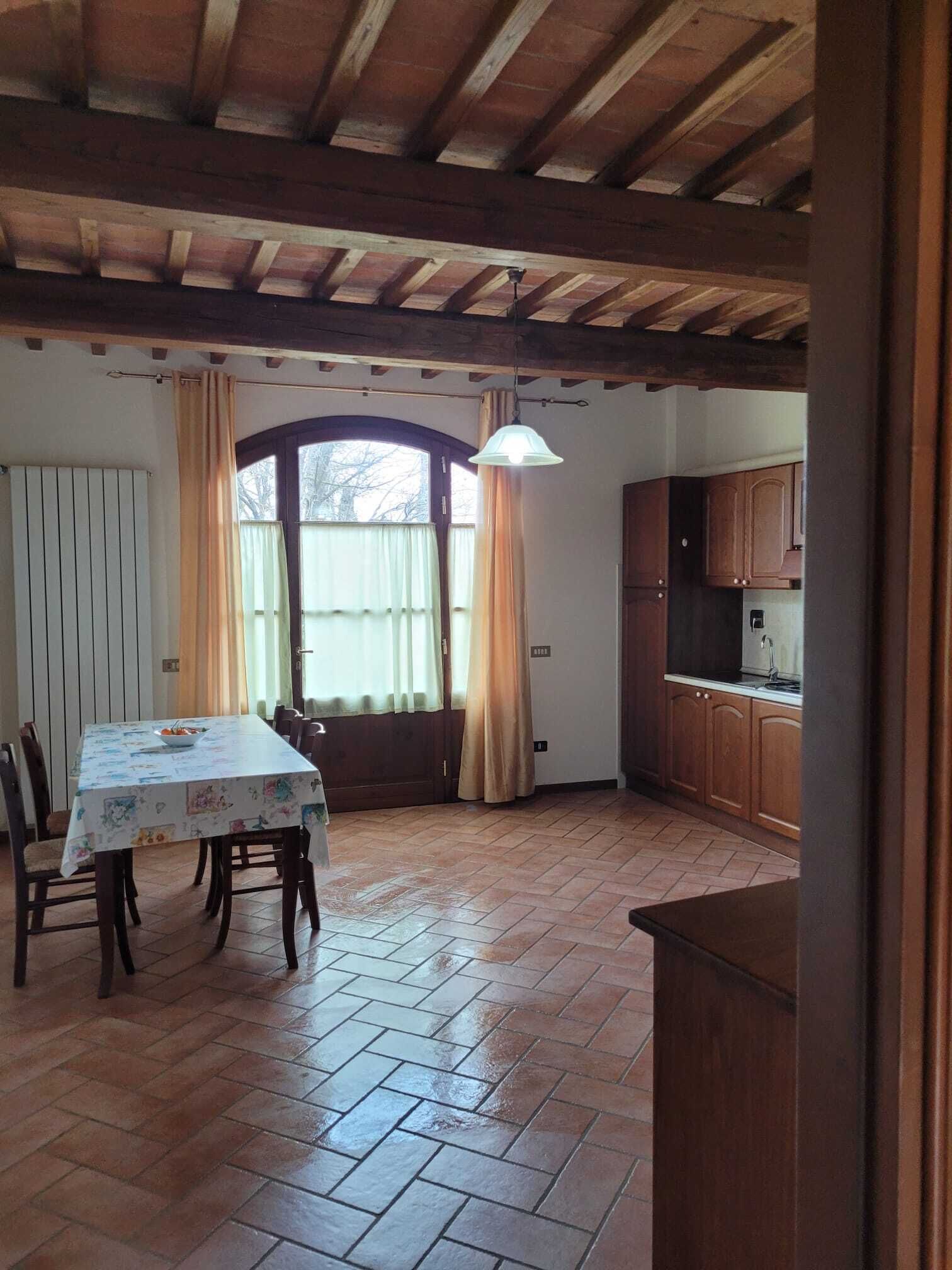 A dining area with a table covered in a tablecloth, wooden chairs, a window with curtains, and a kitchen area with wooden cabinets.