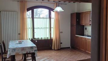 A dining area with a table covered in a tablecloth, wooden chairs, a window with curtains, and a kitchen area with wooden cabinets.