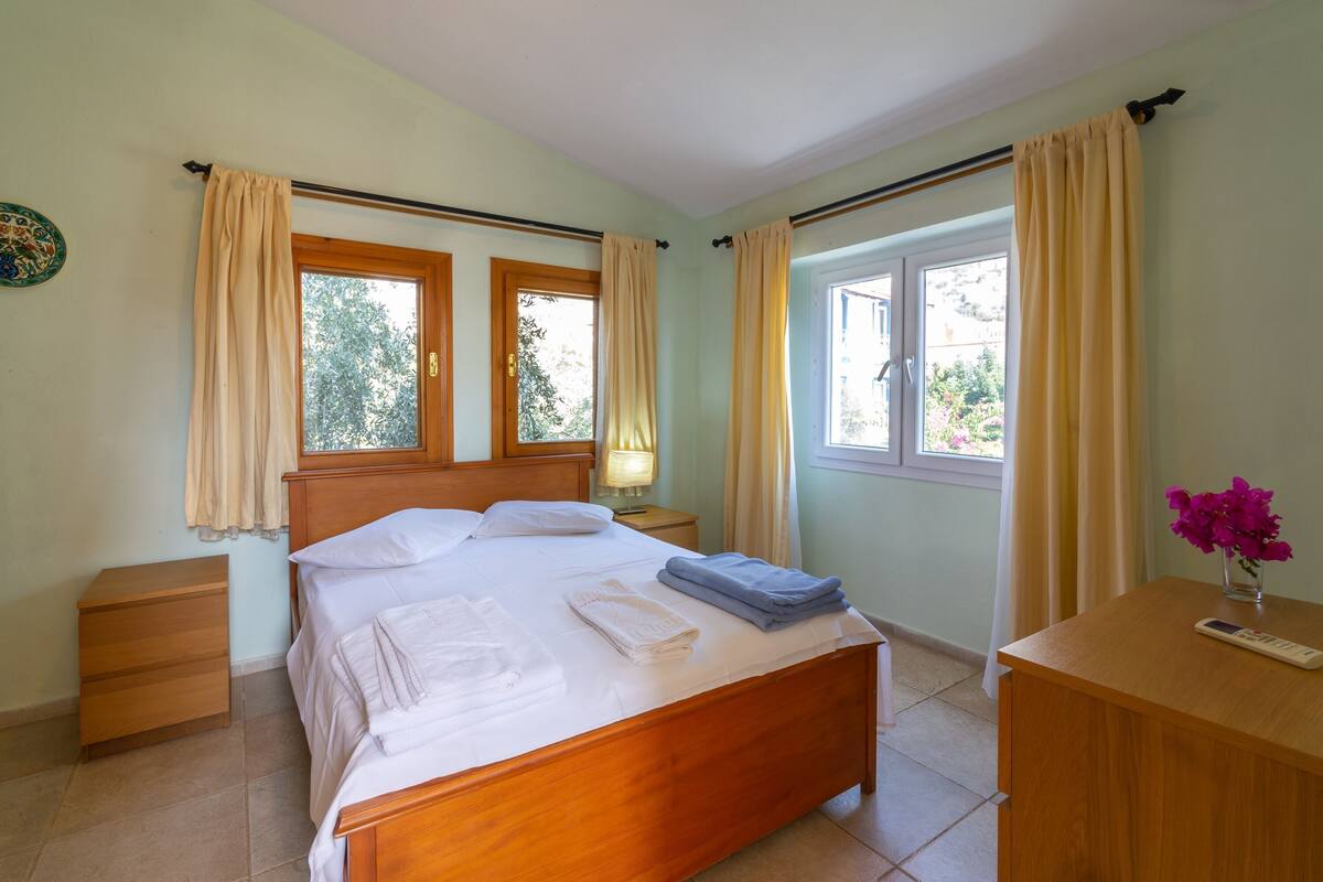A bedroom with a wooden bed, two windows with curtains, a bedside table, a nightstand, and a small table with a vase of flowers.