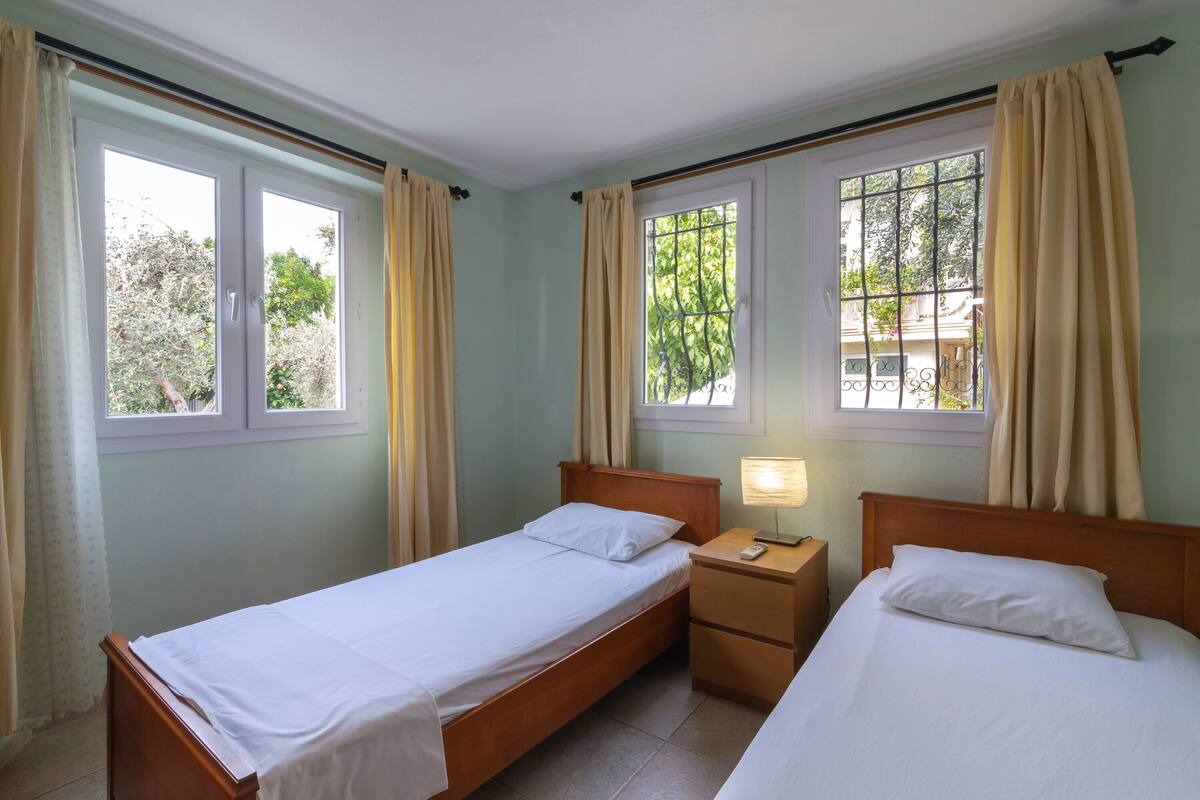 A room with two single beds, a bedside table with a lamp, and windows with curtains.