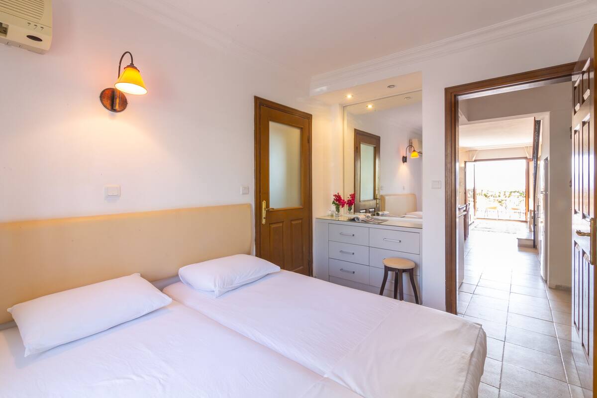 A hotel room with two beds, a wooden door, a bathroom with a mirror, and a view of the outdoor area.