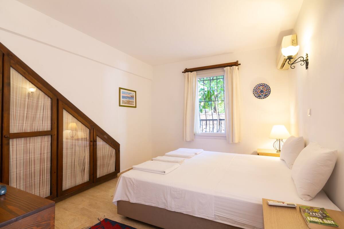 A bedroom with a bed, a nightstand, a lamp, a window with curtains, a picture on the wall, and a wooden desk.