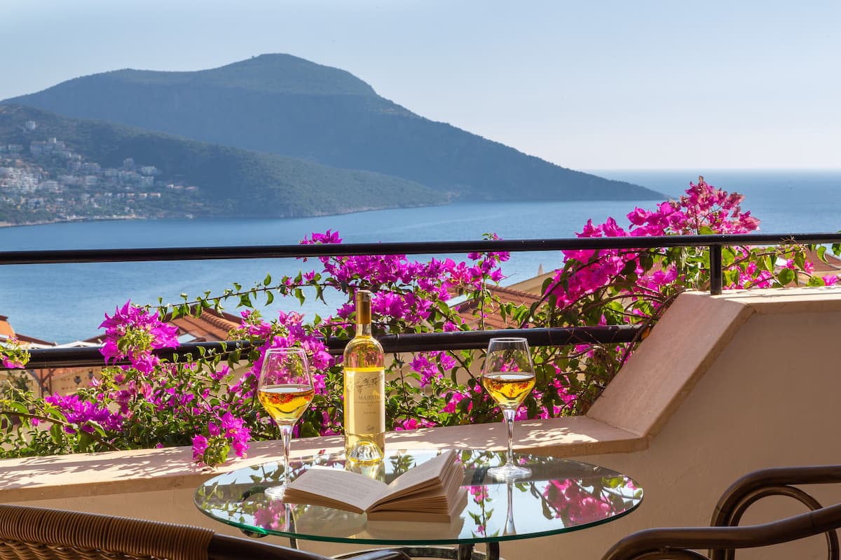 A balcony with a glass table, two wine glasses, a bottle of wine, and a book.