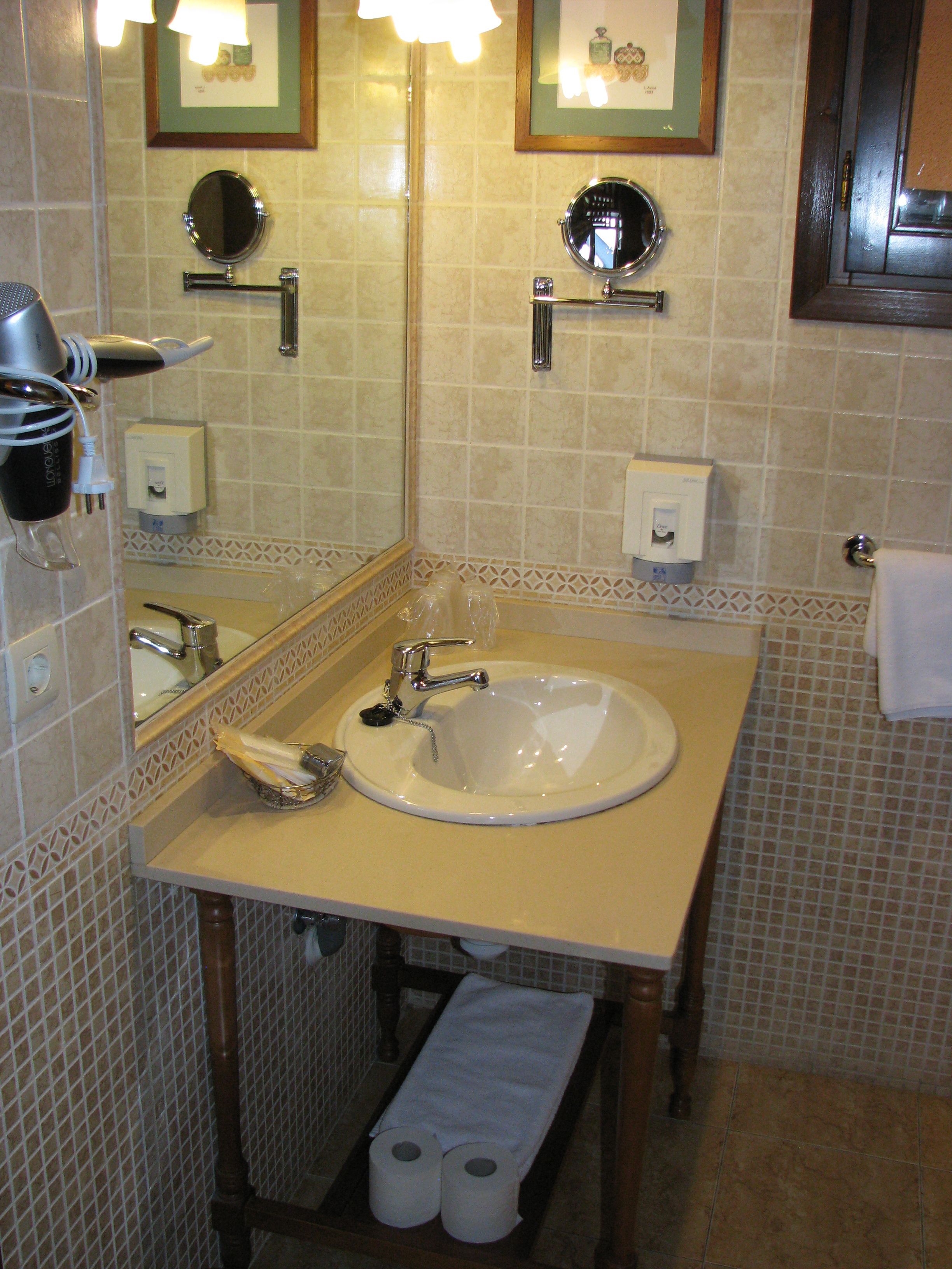 Un baño con lavamanos, espejo, toallero y dispensador de papel higiénico.
