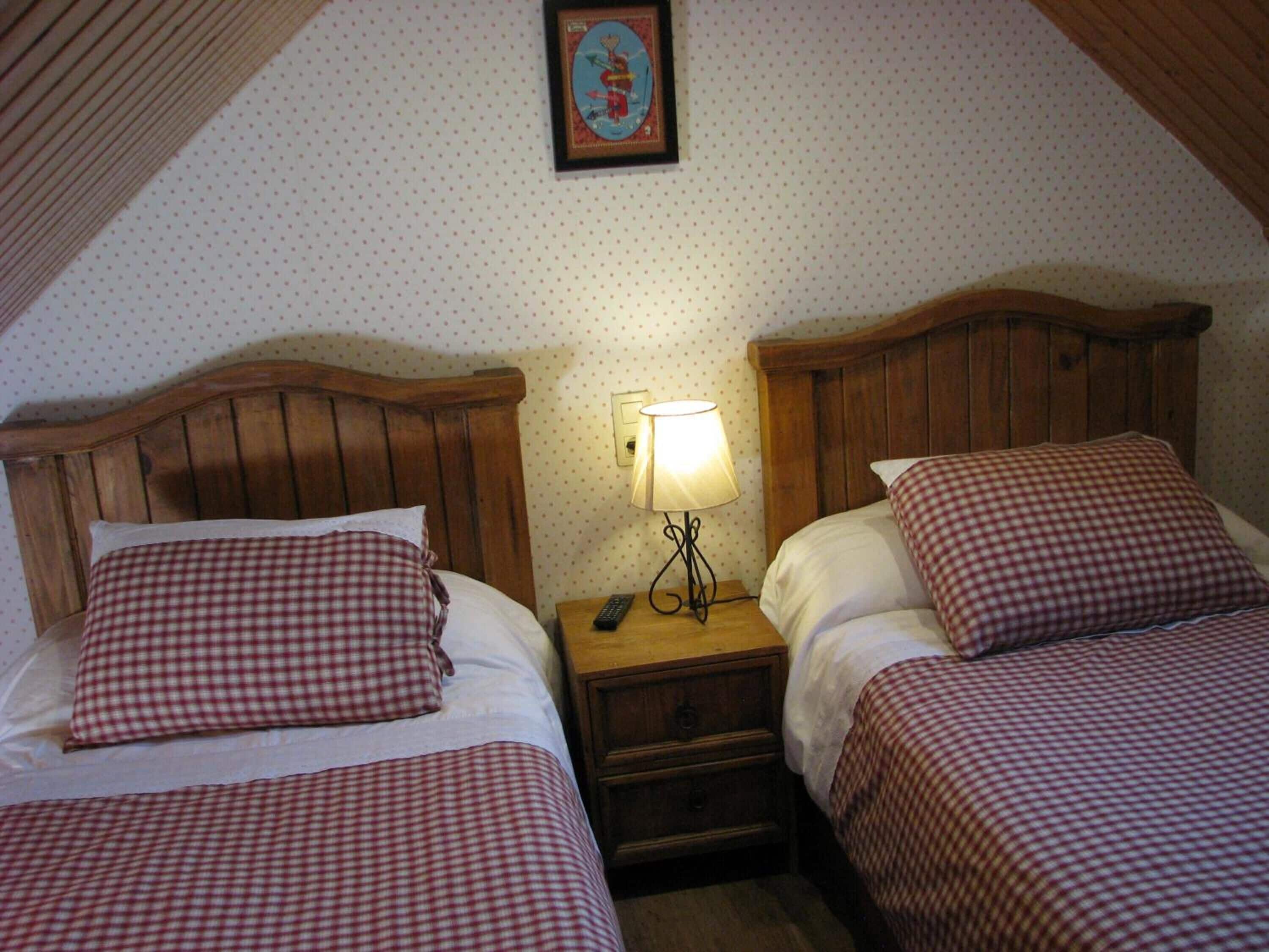 A room with two single beds, each with a checkered bedspread and matching pillows. There is a nightstand with a lamp between the beds. The room has a sloped ceiling and a framed picture on the wall.