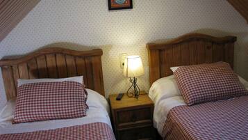 A room with two single beds, each with a checkered bedspread and matching pillows. There is a nightstand with a lamp between the beds. The room has a sloped ceiling and a framed picture on the wall.