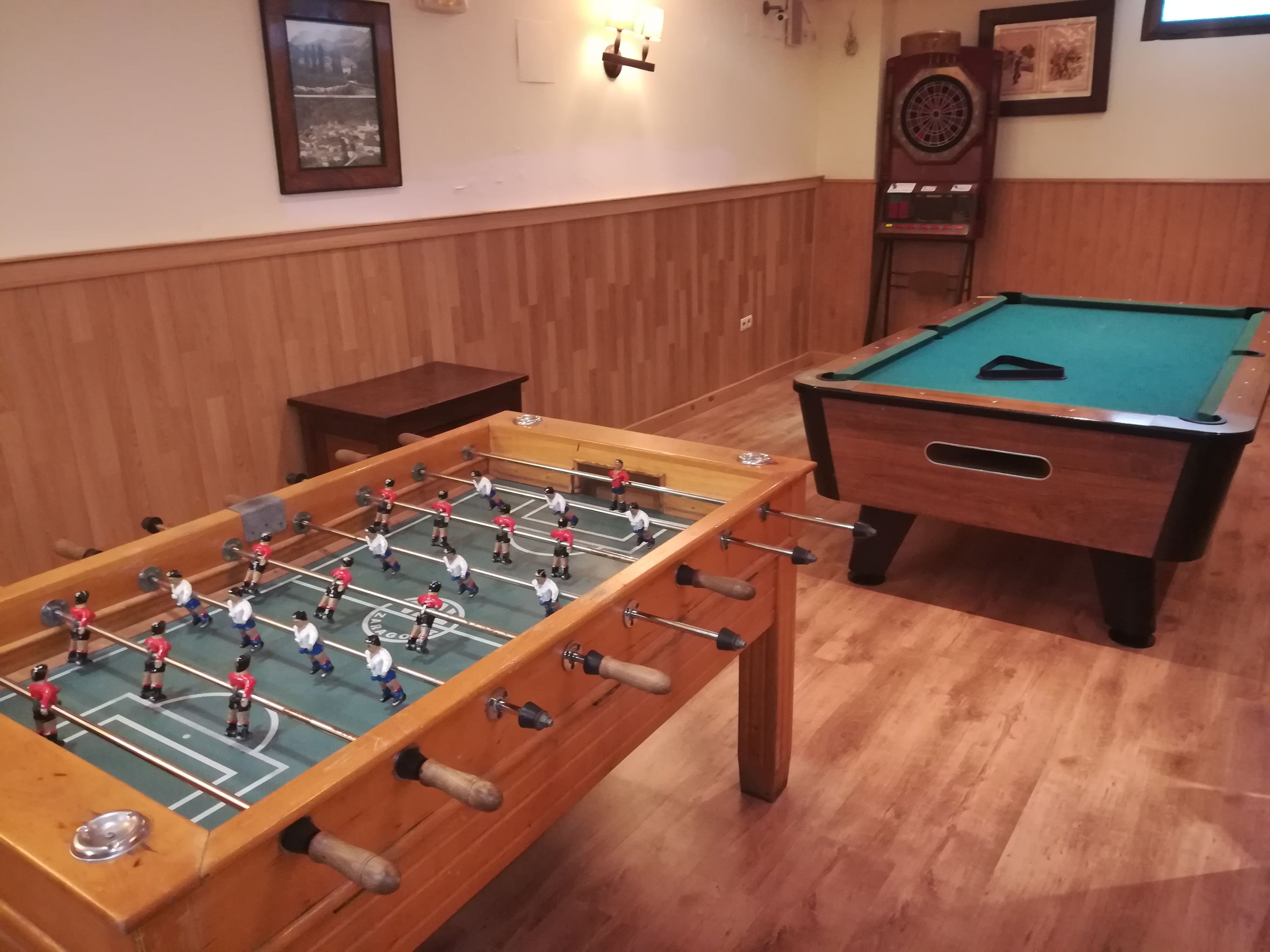 A foosball table and a pool table in a room with wooden paneling and a framed picture on the wall.