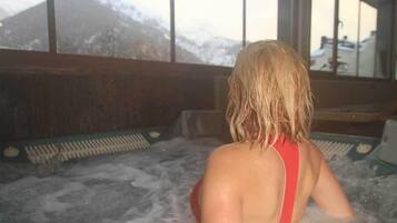 A person in a hot tub with a mountain view through large windows.
