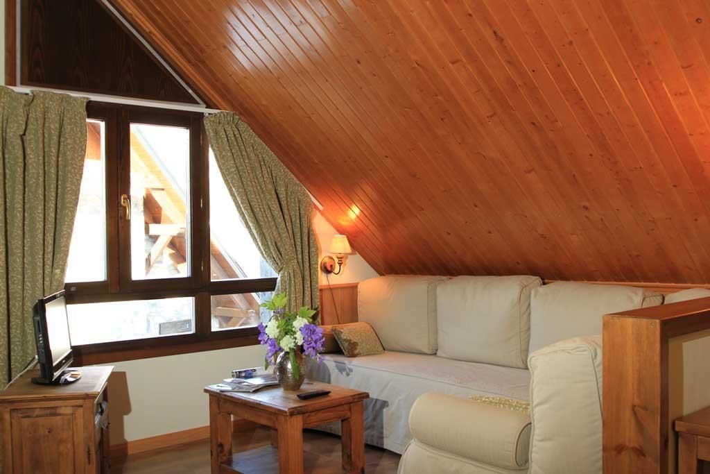 Acogedora habitación en el ático con techo de madera, un sofá, una mesa con un florero y una ventana con cortinas.