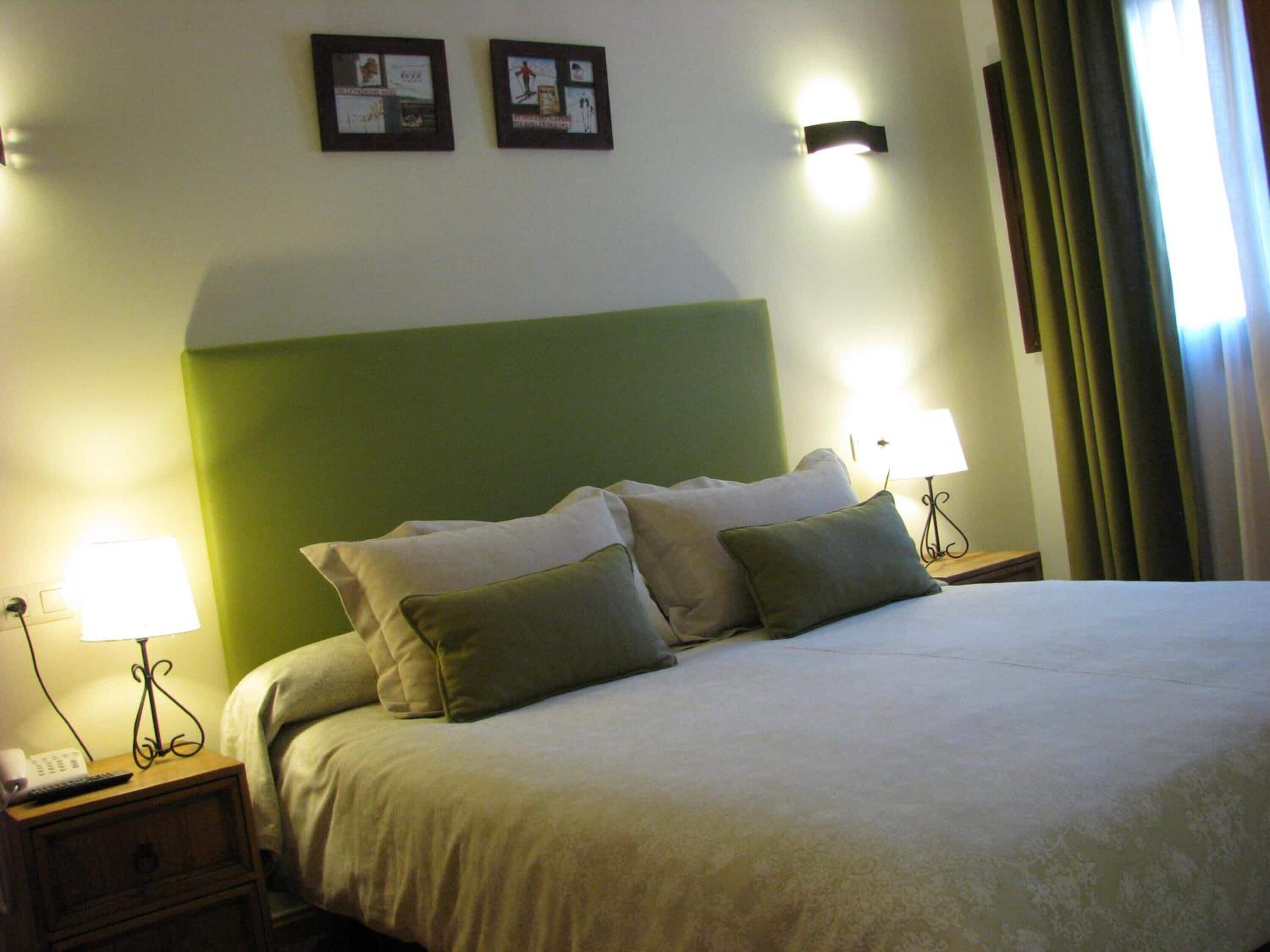A hotel room with a bed, two bedside lamps, a nightstand, a green accent wall, and two framed pictures on the wall.