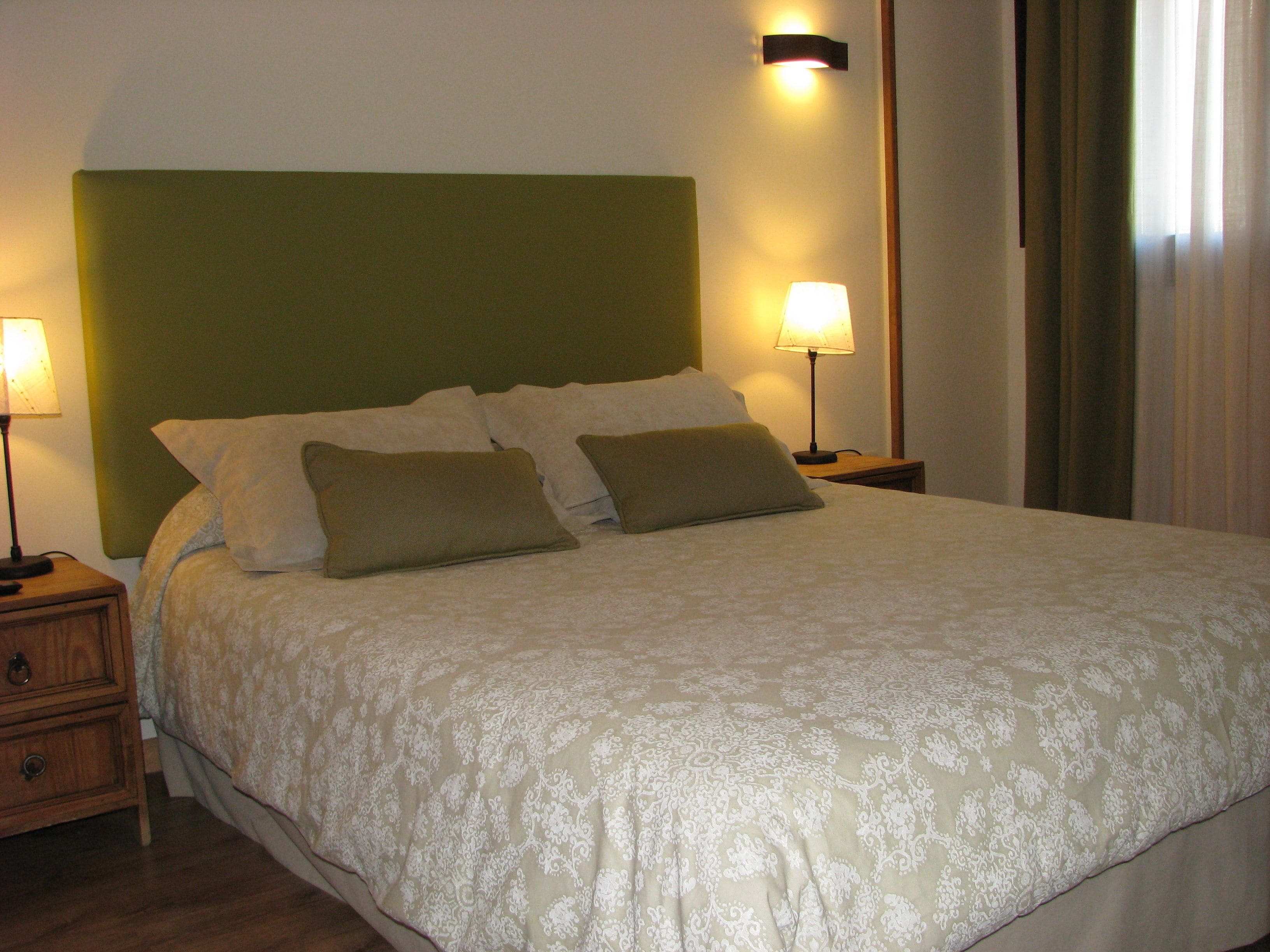 A neatly made bed with a green headboard, two bedside lamps, and a wooden nightstand with a drawer.
