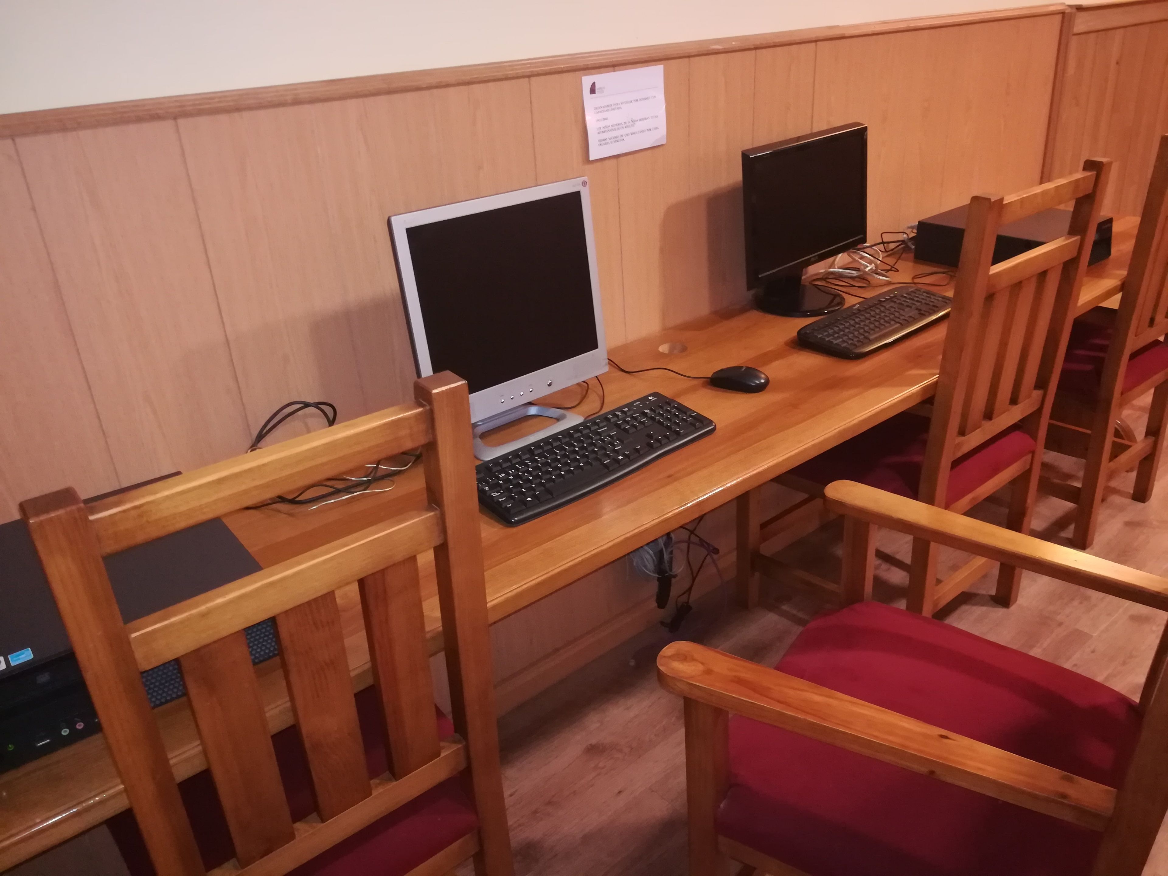 Uma escrivaninha com um monitor, teclado e rato. Há cadeiras de madeira com almofadas vermelhas.