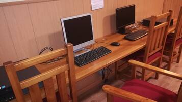 Uma escrivaninha com um monitor, teclado e rato. Há cadeiras de madeira com almofadas vermelhas.