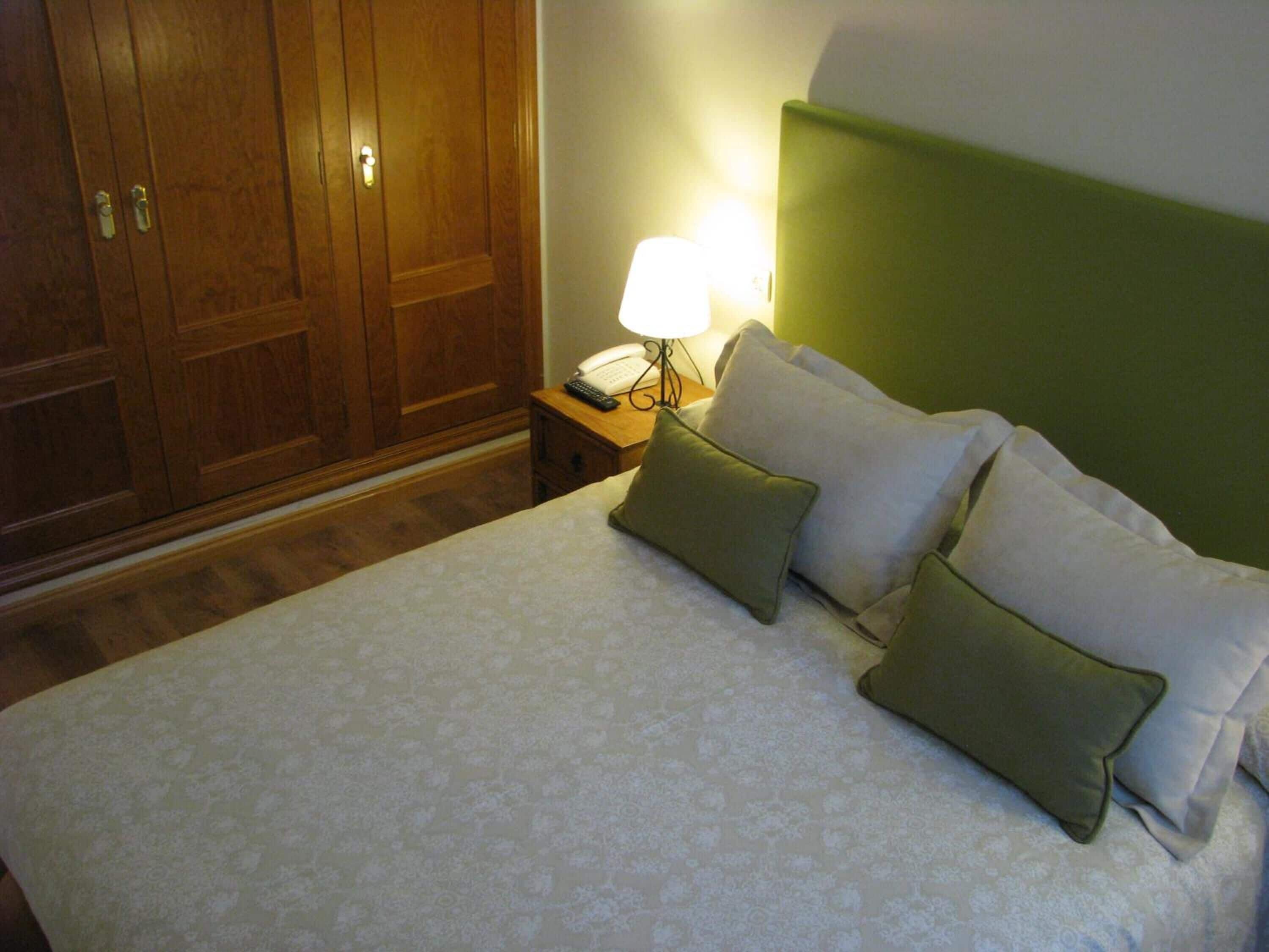 Una cama bien hecha con un edredón estampado, dos almohadas, una lámpara de noche y un teléfono sobre una mesita de noche de madera.
