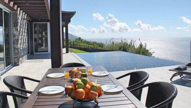 Mesa de comedor con frutero, platos y cubiertos, con vista a una piscina y al océano.