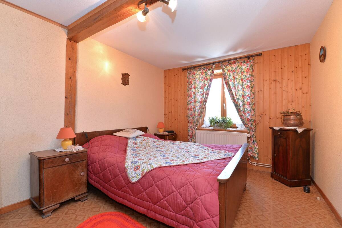 Une chambre avec un plancher en bois, un lit recouvert d’une couette rouge, une table de chevet avec une lampe, une armoire en bois et une fenêtre ornée de rideaux à motifs floraux.